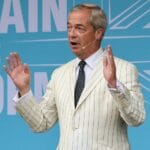 Nigel Farage addressing a Reform UK rally with both hands raised, wearing a cream striped jacket.