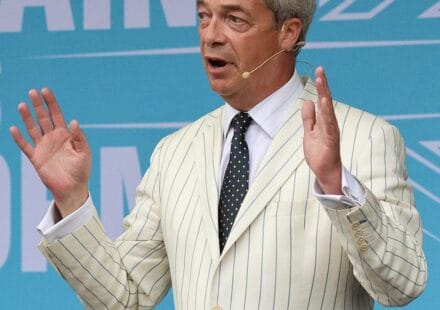 Nigel Farage addressing a Reform UK rally with both hands raised, wearing a cream striped jacket.