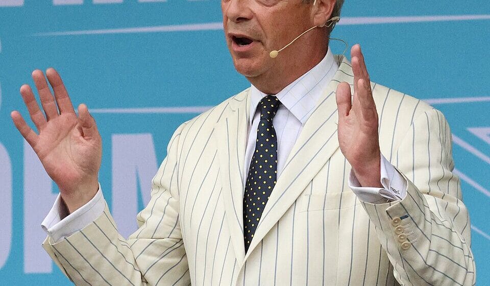 Nigel Farage addressing a Reform UK rally with both hands raised, wearing a cream striped jacket.